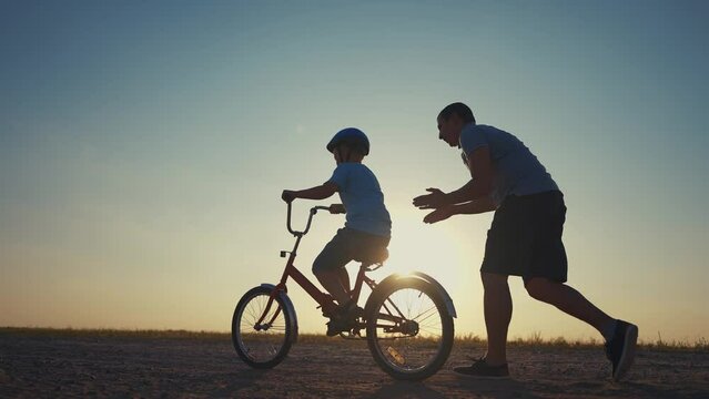 family play in the park. father teaching son to ride a bike. happy family kid dream concept. son learn to ride a bike silhouette. father supporting child sunset son riding bike summer in the park