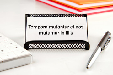 Tempora mutantur et nos mutamur in illis Translated from Latin, it means Times are changing, and we are changing with them. on a white business card next to a calculator, notepad and pen