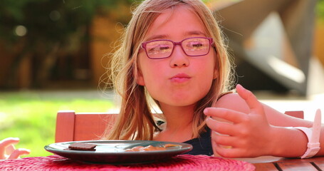 Portrait of little girl with mouth full seated outside at table eating dessert