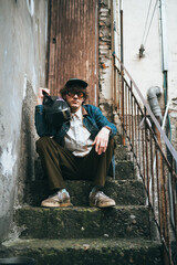 Hip young man with vintage look sits on stone steps with retro cassette player in urban setting, exuding sense of retro fashion and contemplation. 