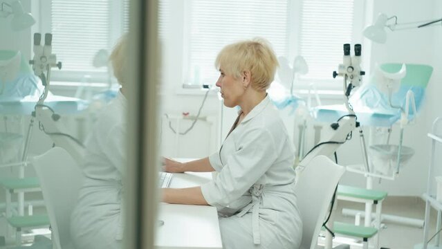 A spectacular female doctor in a medical white coat works at a computer. In the background, her office opens with a gynecological chair and a colposcope. High quality 4k footage