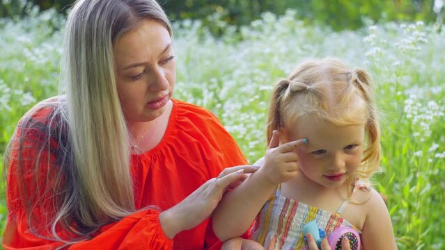 Mom gently paints her daughter's face, but she wants to do it herself. A wonderful time spent in the fresh air among a field of daisies for a joint activity. High quality 4k footage