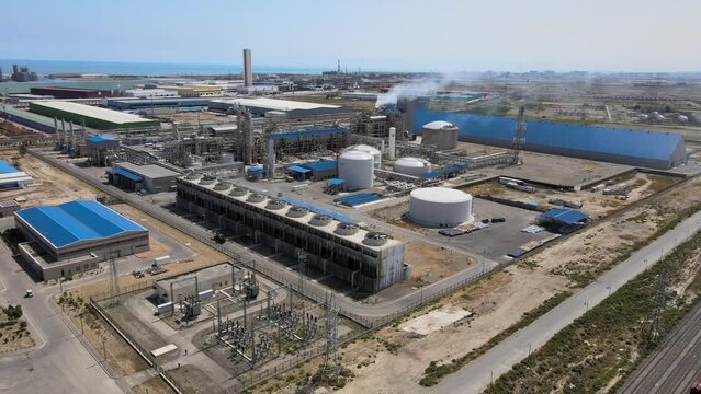 Aerial photography of an industrial area with a variety of buildings and structures. Roads and parking lot can be seen in the foreground and the sea and other industrial structures in the background.