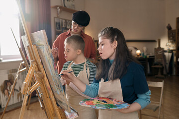 Portrait of two children with disability painting on easels in art class and female teacher assisting