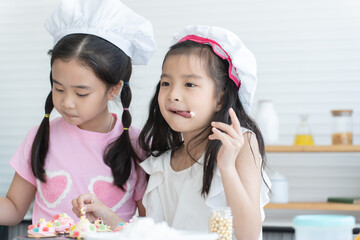 Two cute sisters with chef hat enjoy baking, decorating cupcakes at kitchen at home together. Little children girls have fun tasting cream and homemade cupcake. Happy Asian family. Funny moment