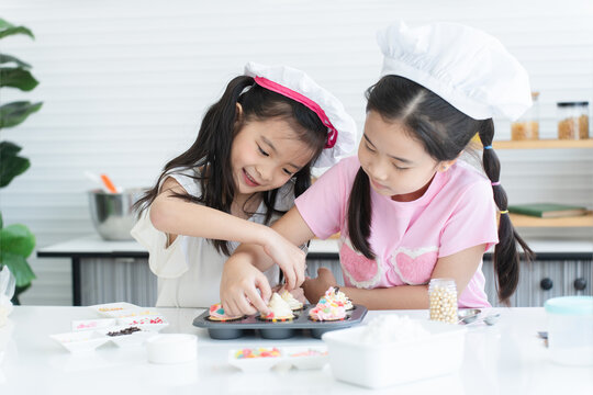 Two cute sisters with chef hat enjoy baking, decorating cupcake with jelly and icing sugar at kitchen at home. Little children girls have fun together. Happy Asian family