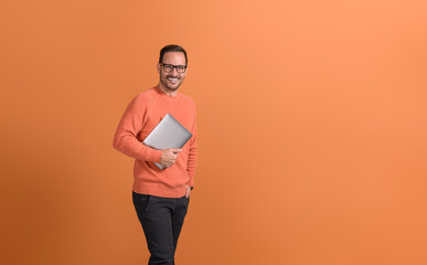 Handsome entrepreneur with hand in pocket holding laptop and smiling at camera on orange background