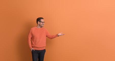 Happy male promoter with hand in pocket looking at empty palm while advertising on orange background