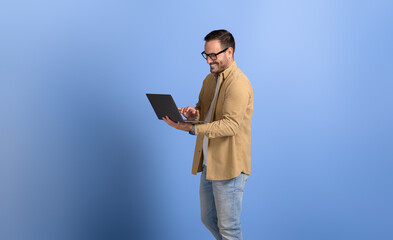 Male manager in eyeglasses smiling and working online over wireless computer against blue background