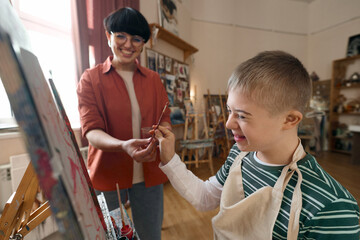 Close up portrait of happy boy with disability standing by easel and holding paintbrush enjoying art class with smiling teacher assisting