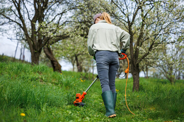 Woman is cutting grass by weed trimmer in garden. Mowing lawn in orchard. Spring gardening