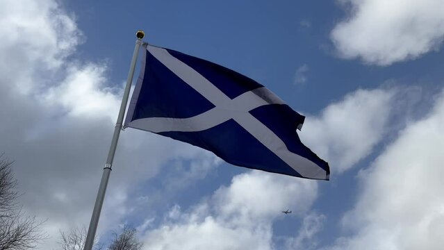Scotland flag (the Saltire), filmed on a sunny, windy day with blue sky and white clouds. A passenger jet passing in the distance.