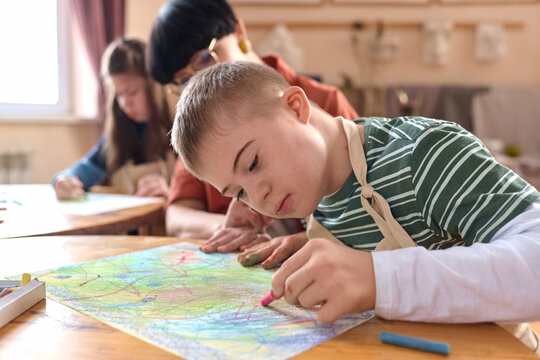 Close up portrait of young boy with Down syndrome painting picture with crayons enjoying art therapy