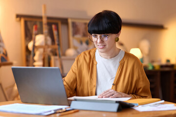 Portrait of smiling female artist using laptop in studio giving online art class and speaking via video chat