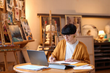 Wide angle view of funky female artist using laptop in art studio and taking notes while managing...