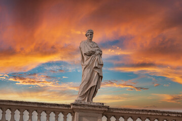 Classical marble statue with draped robe standing under vibrant sunset sky in Vatican. Serene beauty of art and architecture captured in this elegant image.