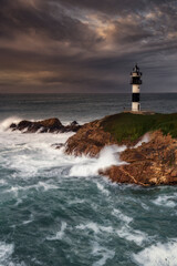 Fototapeta premium Pancha Island Lighthouse in Ribadeo, Lugo, Galicia, on a sunset with many clouds and a dramatic sky with the waves breaking strongly against the coast