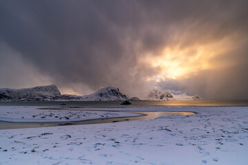 vor dem  Schneesturm spiegelt sich die orange Sonne im Meer, Fluss schl&auml;ngelt sich bei Haukland Beach ins Meer, schneebedeckter Strand