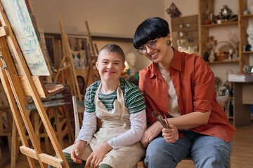 Portrait of smiling boy with Down syndrome enjoying art class with female teacher and looking at camera