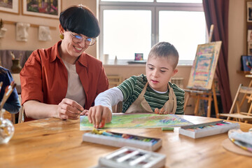 Portrait of young boy with Down syndrome enjoying art class with female teacher assisting and...