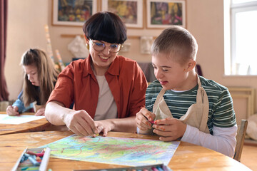 Portrait of smiling boy with disability enjoying art class in studio with mentor assisting