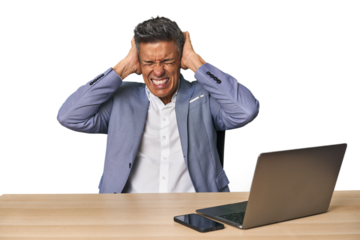 Elegant businessman at desk with laptop covering ears with hands trying not to hear too loud sound.