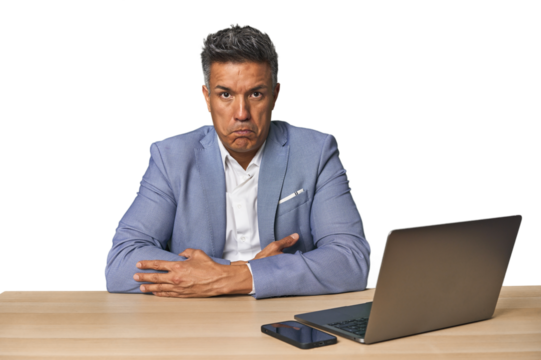 Elegant businessman at desk with laptop blows cheeks, has tired expression. Facial expression concept.