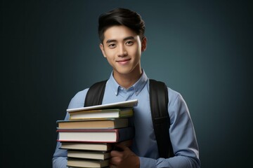Confident young male student carrying a stack of books, wearing a light blue shirt and backpack, isolated on a dark background with copy space.