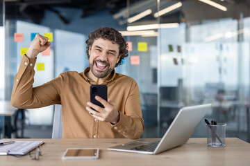 Joyful businessman celebrating a successful deal with a fist pump while looking at his smartphone,...