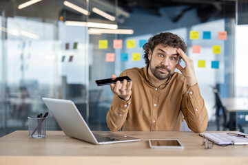 A mature businessman appears confused while using a smartphone in a modern office setting. Problem-solving concept.
