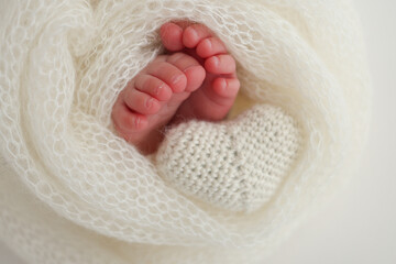 The tiny foot of a newborn baby. Soft feet of a new born in a white wool blanket. Close up of toes, heels and feet of a newborn. Knitted white heart in the legs of a baby. Macro photography