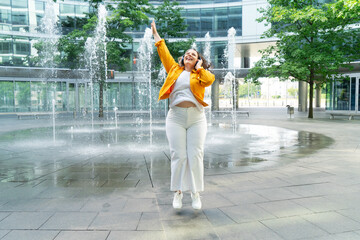 Happy curvy woman in city, plus size brunette lady with curly gray hair jumping emotionally in front of the fountains in modern city