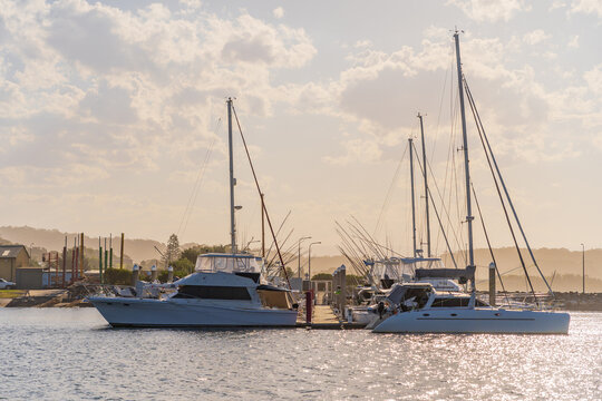 Tall masts on a row of yachts moored at a coastal marine with hazy clouds over sparkling water