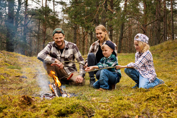 family with two children frying sausages over a bonfire while camping in forest. family time, nature adventure - 779963932
