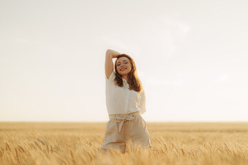 Happy young woman in a white shirt standing in a wheat field at sunset, experiencing the beauty of...