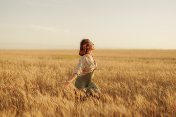 Carefree adult woman with arms stretched out standing in a vast wheat field at sunset, feeling...