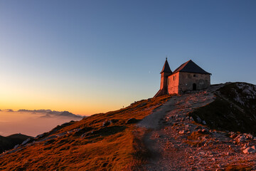 Scenic sunrise view of chapel Maria am Stein on top of mountain peak Dobratsch, Villacher Alps, Austria, Europe. Looking at Julian and Karawanks mountain range. Golden morning hour tranquil atmosphere