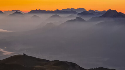 Panoramic sunrise view from Dobratsch on Julian Alps and Karawanks in Austria, Europe. Silhouette of endless mountain ranges with orange and pink sky. Jagged sharp peaks and valleys. Cottage on hill