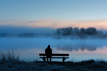 A man sits on a bench by a lake, looking out at the water - Powered by Adobe