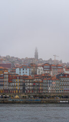 Douro River and promenade of Porto with the Clérigos Church standing out among the houses and roofs of the historic neighborhood covered by fog and rain clouds on a romantic day.