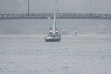Tourists and friends celebrating and having a good time on a recreational sailboat sailing along the Douro River passing under the Don Luis I Bridge in Porto on a very foggy and rainy day.