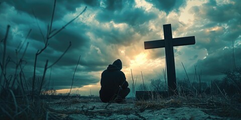 A person is kneeling in the grass in front of a cross. The sky is cloudy and the sun is setting