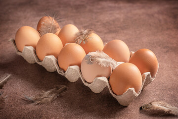 Group of raw fresh whole chicken eggs in carton box or container made of recycled paper with hen feathers on dark brown table used as ingredient in culinary full of protein for healthy breakfast