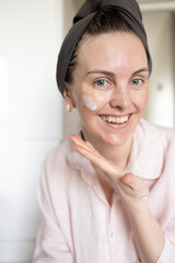Woman in turban after washing hair looking at camera, smiling and showing cosmetic cream smear on face. Concept of morning skincare routine, anti-age care, hydration, no retouch