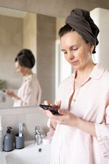 Woman in turban after washing hair scrolling or typing on smartphone in bathroom at home