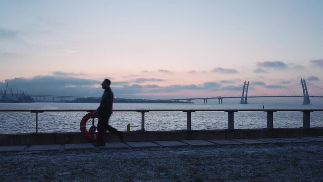 Man walks briskly along promenade at dusk, large suspension bridge in background. Silhouette strides past lifebuoy, evening sky painted with hues of pink and blue. Themes: solitude, urban evening