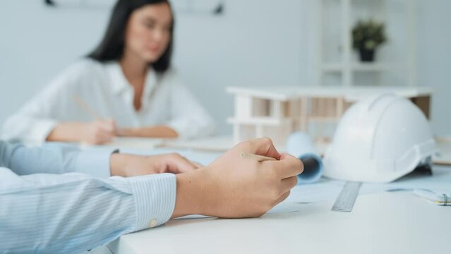 Professional architect engineer drawing blueprint and working together with young beautiful caucasian coworker on meeting table with safety helmet, house model, blueprint scatter around. Immaculate.