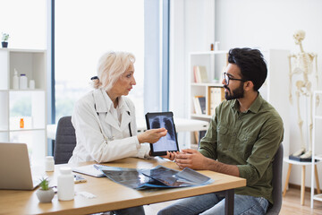 Obraz premium Mindful gray-haired female in lab coat holding tablet with MRI scan while brunette guy sitting next to her at desk. Family doctor examining diagnostic test results while developing treatment plan.