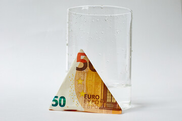 Economy. Increasing the cost of water is a concept. 50 euro banknote folded in triangle next to glass filled with quarter of water