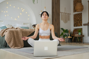 Yoga mindfulness meditation online. Woman practicing yoga with online lessons in laptop at home. Woman sitting in lotus pose meditating relaxing indoor. Girl doing breathing practice. Yoga at home